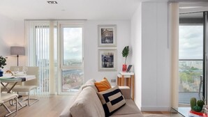 Una sala moderna con un sofá, una mesa de comedor con sillas y una mesa de centro de vidrio. Hay dos ventanas con vistas a la ciudad y una planta pequeña sobre una mesita auxiliar.