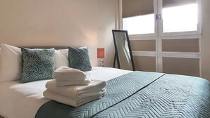 Una camera d'hotel con un letto, cuscini, una pila di asciugamani piegati, una finestra con tende e uno specchio fissato alla parete.
