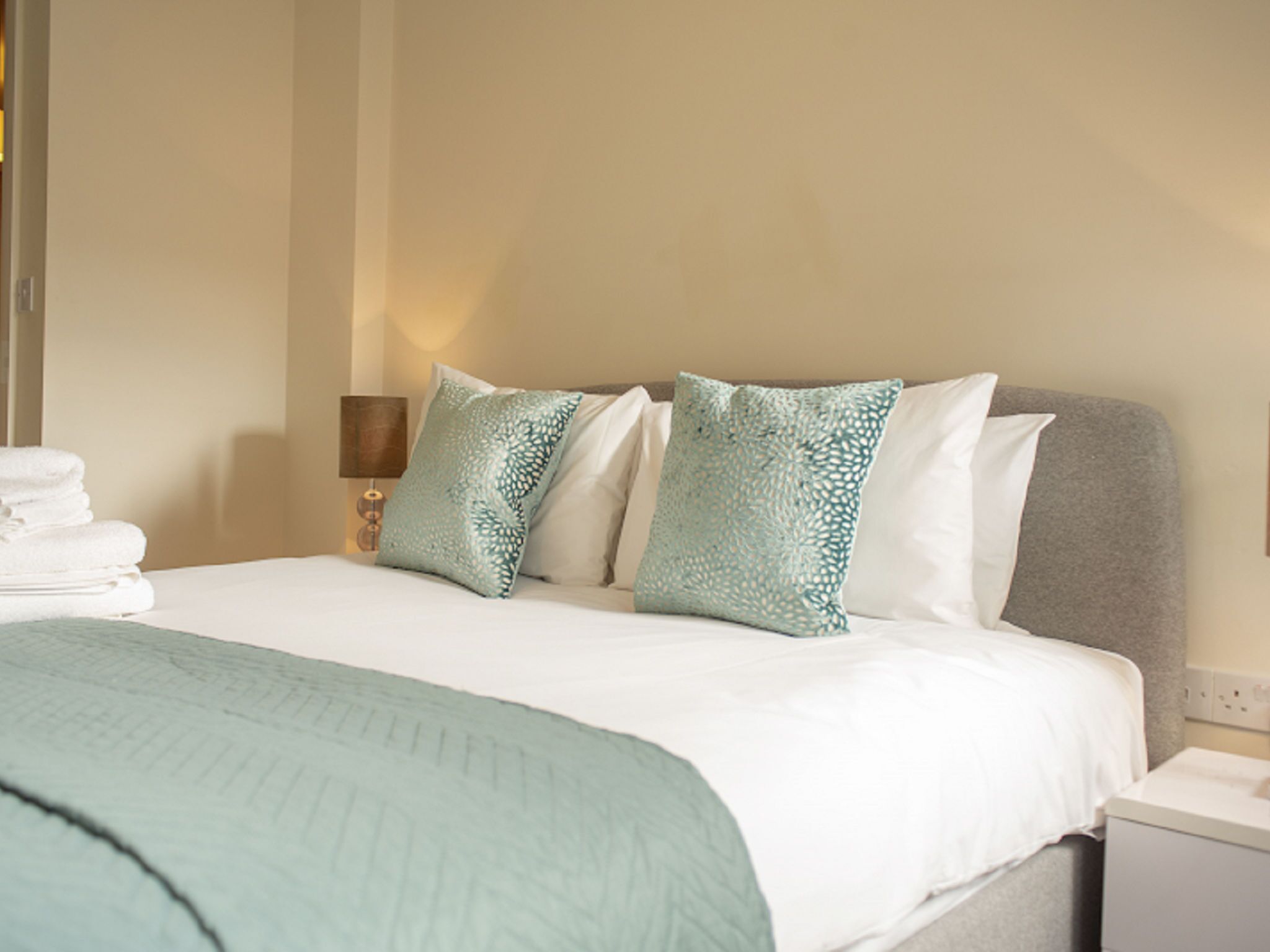 A neatly made bed with a white comforter and two patterned pillows, a teal throw blanket, and a bedside lamp on a nightstand.