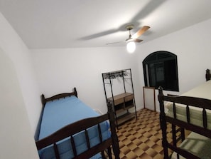 Een kleine, smalle kamer met een enkel bed, een plafondventilator, een raam en een klein tafeltje.