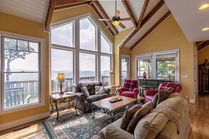Ein geräumiges Wohnzimmer mit großen Fenstern, einem Deckenventilator und Blick ins Freie.