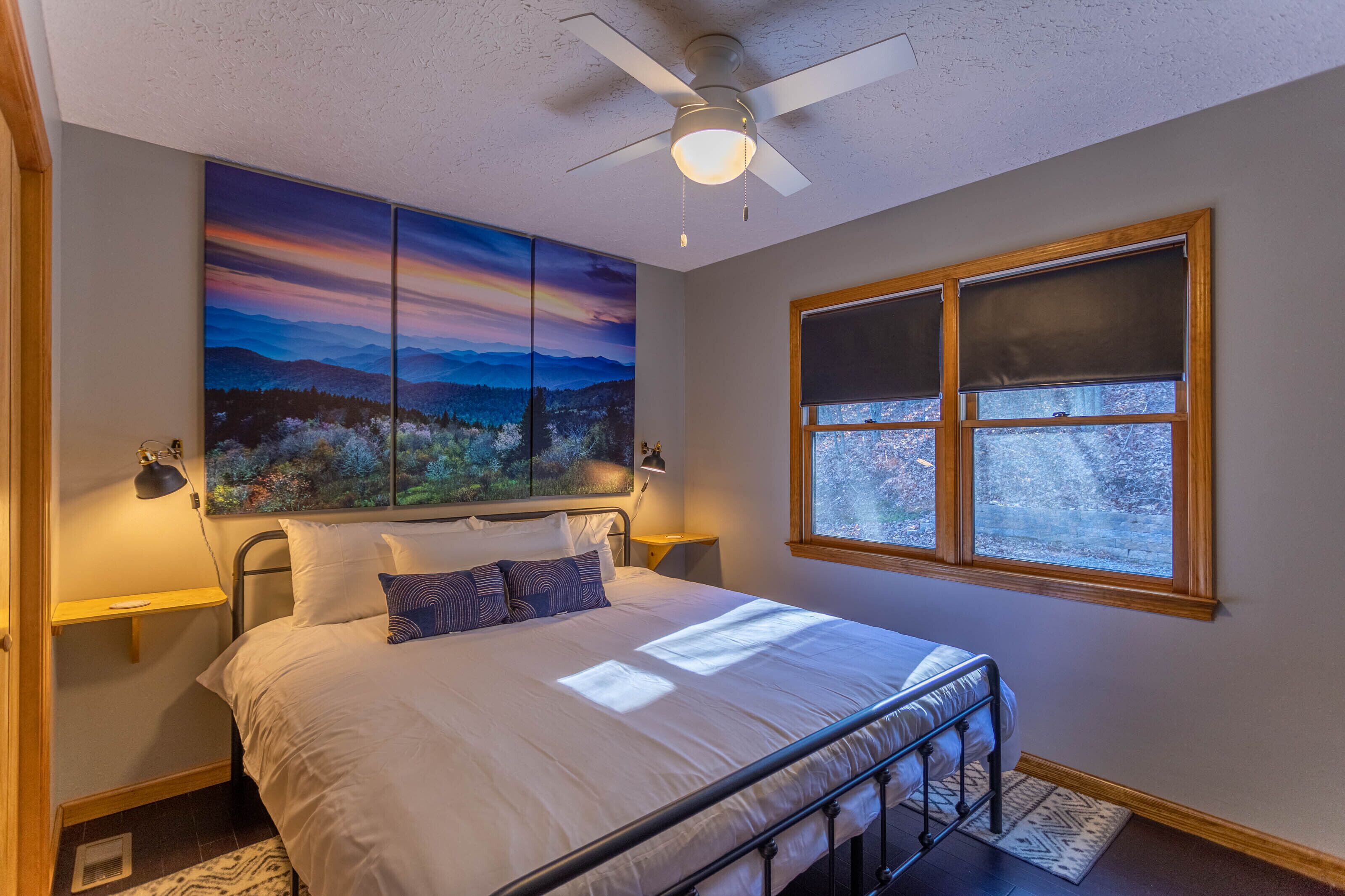 A bedroom with a large bed, a ceiling fan, a window with blinds, and a wall-mounted painting.