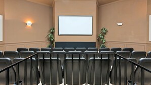 Una sala de conferencias con hileras de sillas negras, una pantalla grande al frente y dos luces fijadas a la pared.