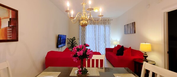 Un coin repas comprenant une table, des chaises, un lustre, un canapé rouge, un téléviseur et une plante.