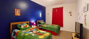 Une chambre avec deux lits, chacun agrémenté de draps à l’effigie de Mickey Mouse, un rideau rouge et un téléviseur posé sur un meuble.