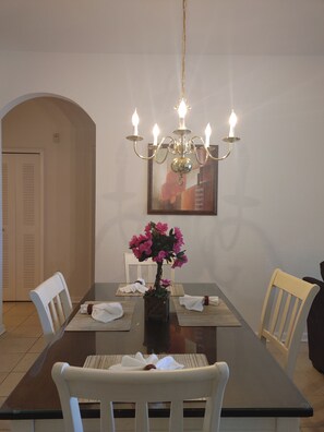 Un coin repas comprenant une table rectangulaire, des chaises blanches, un lustre et un vase rempli de fleurs roses.