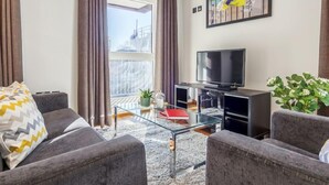 Una sala de estar moderna con un sofá, una mesa de centro de vidrio, un televisor de pantalla plana y un ventanal con cortinas.