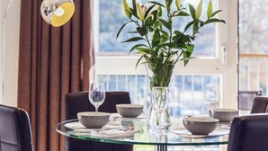 Una mesa de comedor con cristalería y fuentes, un florero transparente con plantas verdes, y una lámpara de pie moderna.
