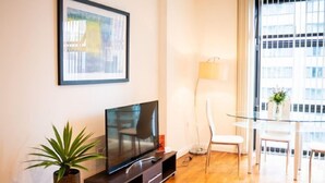 Una sala moderna con un televisor de pantalla plana sobre un soporte de madera, un cuadro en la pared, una lámpara de pie, una mesa de comedor con sillas y una planta en maceta.