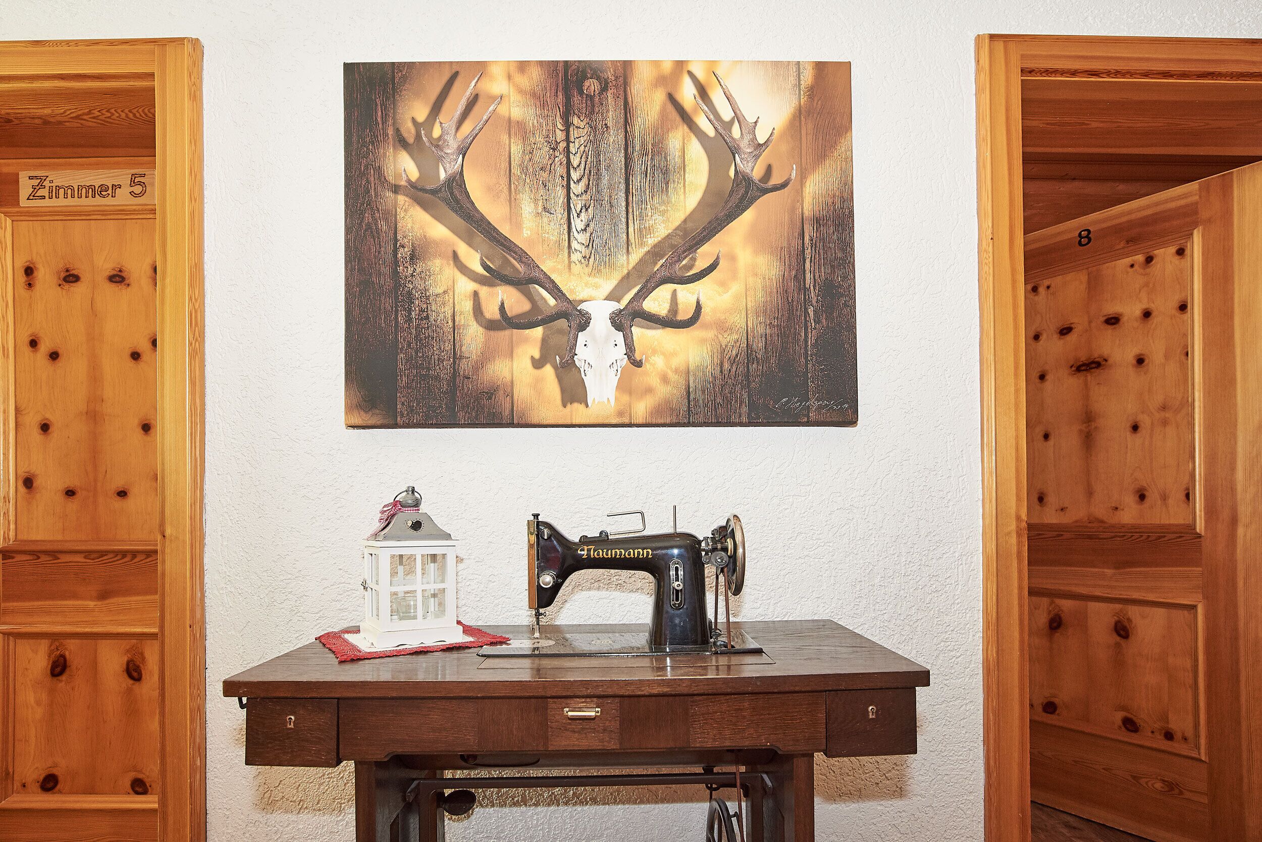 A wooden desk with a vintage sewing machine, a small white lantern, and a deer skull painting on the wall.