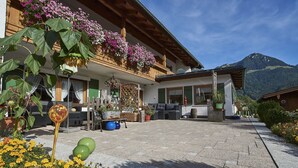 En traditionell chalet med en stenbelagd uteplats, en träbalkong full av blommor och en bergskedja som bakgrund.