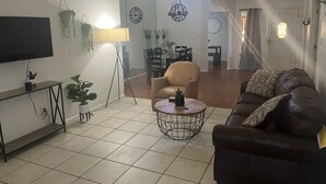 Uma sala de estar com sofá, mesa de centro, TV e um espaço de jantar ao fundo.
