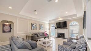 A spacious living room with a fireplace, grey sofas, a patterned armchair, and artwork on the walls. - Relax in our spacious villa, just a quick ride away to Grapevine Malls & Airport (Highland Village)