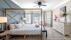 A bedroom with a four-poster bed, a ceiling fan, a bedside table with a lamp, a bench, a dresser, and a window with curtains. - Relax in our spacious villa, just a quick ride away to Grapevine Malls & Airport (Highland Village)