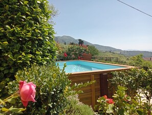 Una piscina rectangular con cubierta de madera, rodeada de abundante vegetación y plantas con flores, con un paisaje montañoso al fondo.