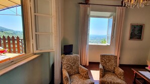 Una habitación con un ventanal grande que ofrece vistas a un paisaje verde, dos sillas con estampado floral, una mesa con un jarrón de flores y un cuadro en la pared.
