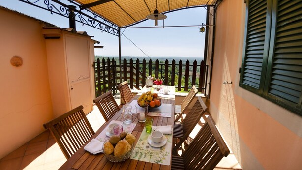 Una mesa de madera dispuesta con frutero, platos y vasos, rodeada de sillas en un balcón con vistas.