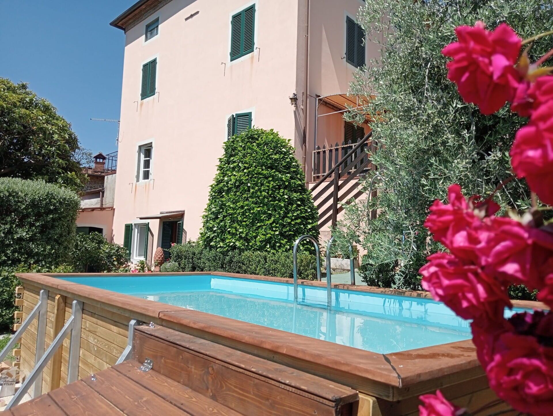 A swimming pool with a wooden deck, surrounded by greenery and a building with multiple windows.