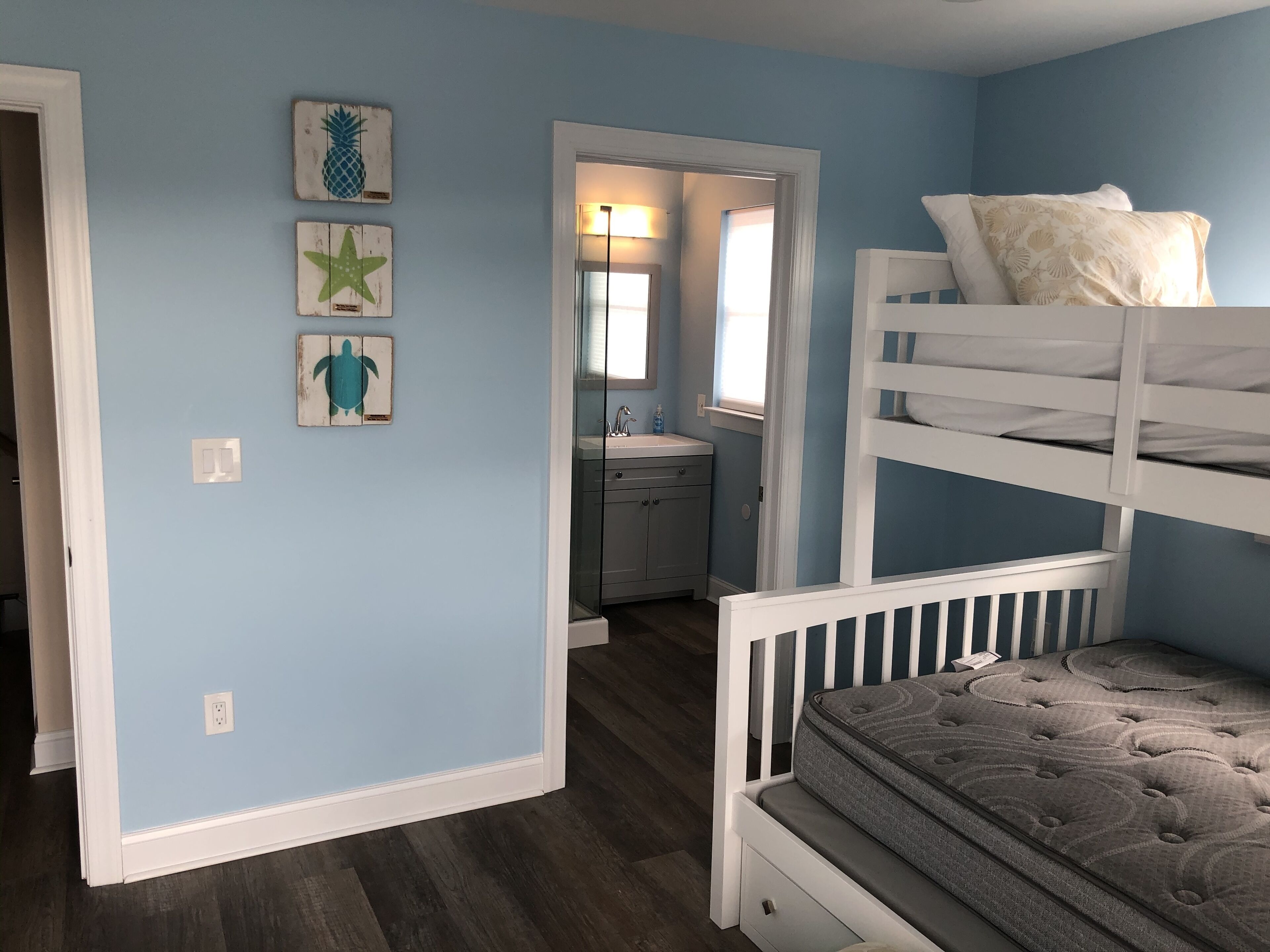 A child's bunk bed with a grey mattress and white headboard, a small bathroom with a sink and mirror, and three wall art pieces depicting a pineapple, a starfish, and a turtle.