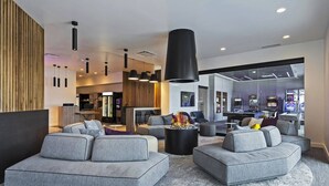 A modern lobby with grey sofas, a central fire feature, and a bar area in the background.