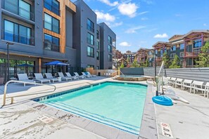 A swimming pool with lounge chairs and a water slide, surrounded by modern buildings with balconies. - Canyon Haus 223 (Park City)