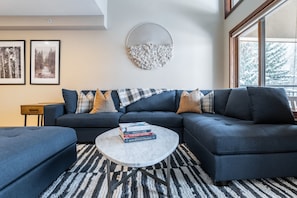 Ein modernes Wohnzimmer mit einem dunkelblauen Ecksofa, einem runden Couchtisch mit Büchern, einem gemusterten Teppich und gerahmten Bildern an der Wand.