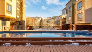A swimming pool with wooden deck, stone and wood buildings, and a mountain backdrop. - Cloud 9 by AvantStay | Located at the Base of Vail Ski Resort w/ Fire Pit + BBQ (Vail)