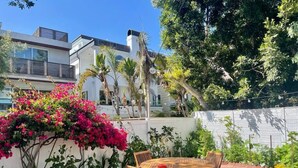 A patio with a round wooden table and chairs, surrounded by lush greenery and flowering plants. - King 1BR, Steps to the Beach on Walk St with Patio and Garden, Prime Location (Venice)