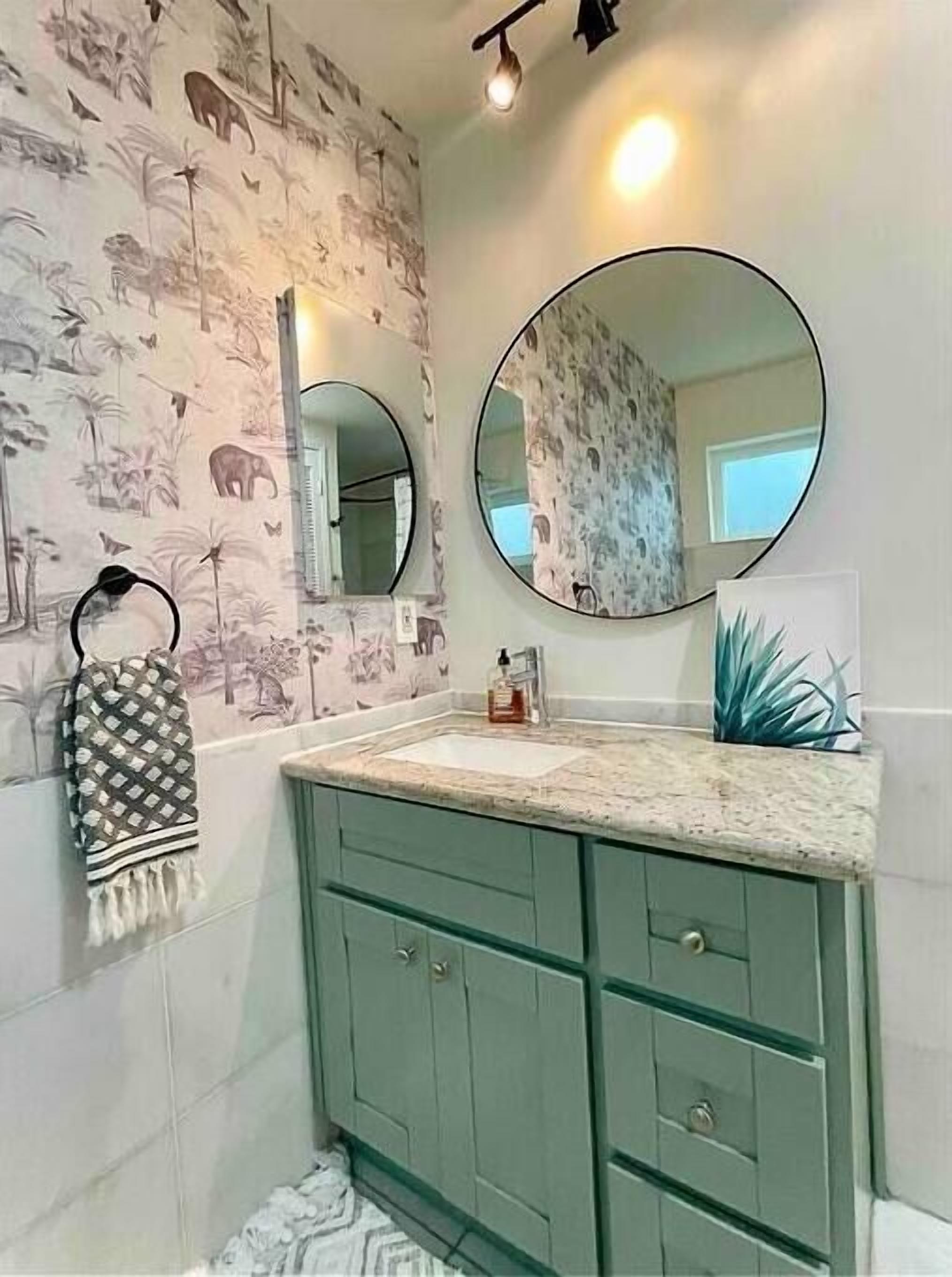 A bathroom with a patterned wallpaper, a round mirror, a sink, and green cabinets.