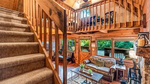 A wooden interior with a staircase, a balcony, and a seating area with a view of greenery.
