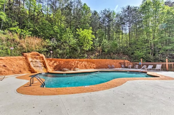 Une piscine dotée d’une terrasse en bois et de chaises longues, entourée d’une zone boisée.