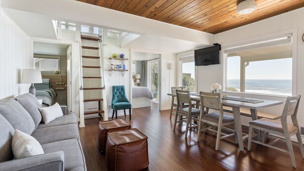 A modern living room with a sofa, ottomans, a teal chair, a dining table with chairs, a ladder, and a view of the ocean. - Oceanfront- Sea Gem Cottage (South Beach)