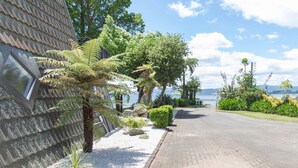 Un camí pavimentat que condueix a un edifici amb teulada d'estries, envoltat de plantes i arbres diversos, amb una massa d'aigua al fons.
