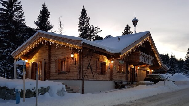 A wooden cabin with a snow-covered roof, string lights, and a sign indicating it is a restaurant.