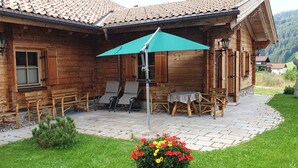 A wooden cabin with a patio area featuring a table and chairs, surrounded by a stone pathway and a flower bed.