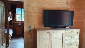 A wooden cabin room with a television on a cabinet, a bed, and a window with a view of greenery.