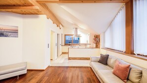 A modern interior with a wooden ceiling, a sofa with cushions, a kitchen area, and a large window. - Apartment "Huck" with Mountain View, Balcony & Wi-Fi (Karneid)