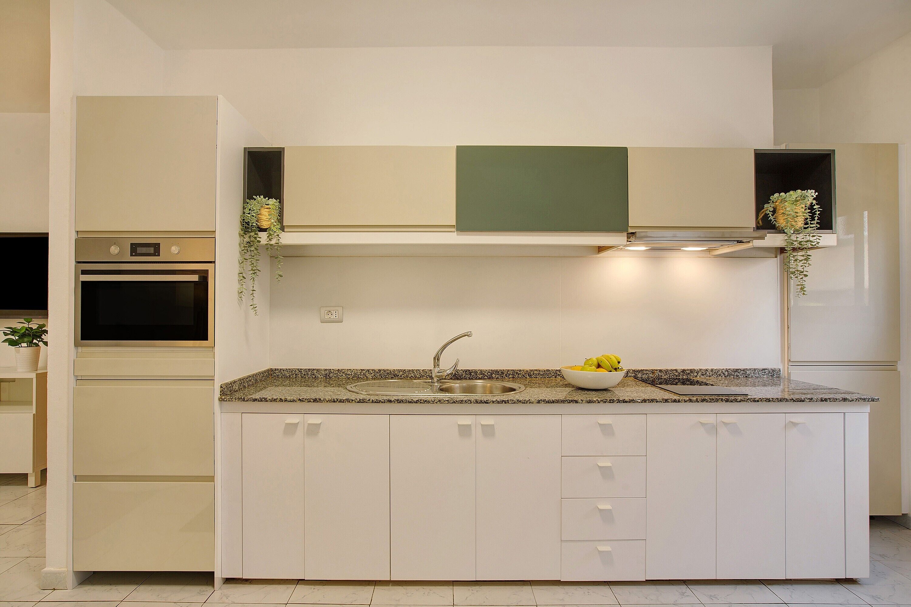 Una cuina moderna amb armaris blancs, taulell de granat i forn integrat, i un rentaplat de stainless steel.