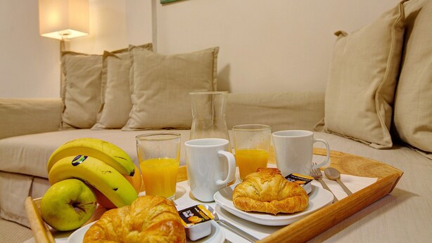Frühstückswagen mit Croissants, Bananen, Saft und Kaffee auf einem Bett mit Kissen und einer Lampe.