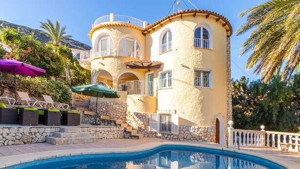 Vista da piscina de um edifício em estilo mediterrâneo, com escadaria de pedra, área de estar externa e piscina azul cristalina.