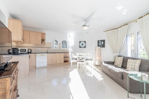 A modern kitchen with wooden cabinets, a dining area with a white table and chairs, and a living area with a sofa and curtains.