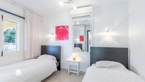Quarto de hotel com duas camas de solteiro, uma pequena mesa de cabeceira com abajur, um espelho grande e uma janela com cortinas.