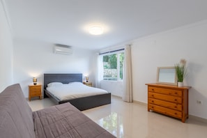 A bedroom with a bed, a dresser, a nightstand, a window with curtains, and an air conditioning unit.