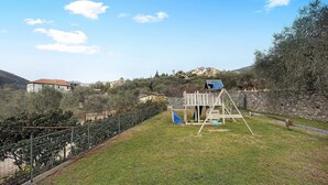 Ein eingezäunter Garten mit einer hölzernen Spielanlage, einer Steinmauer und Häusern im Hintergrund.
