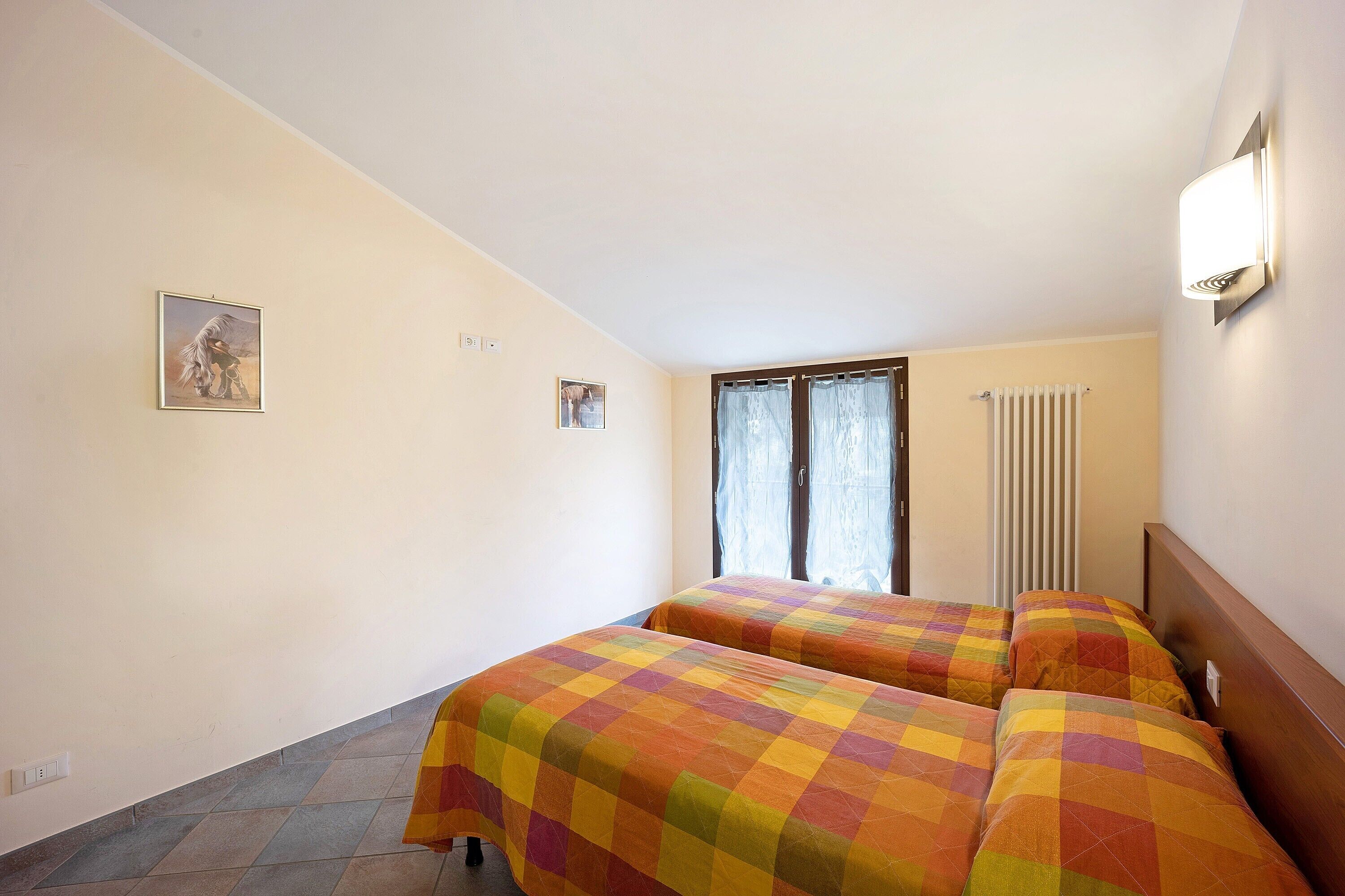 A room with two beds, a radiator, a window with curtains, and a wall-mounted light fixture.