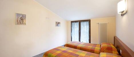 A room with two beds, a radiator, a window with curtains, and a wall-mounted light fixture.