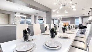 Un comedor moderno con una larga mesa dispuesta para una comida, sillas blancas y un candelabro contemporáneo.