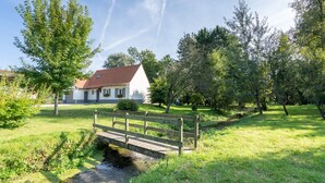 A white building with a red roof, a wooden bridge over a stream, and surrounding greenery. - Fully self-contained cottage (Inxent)