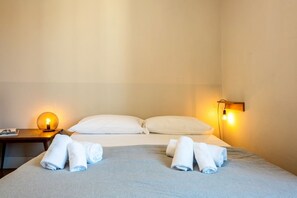 A hotel room with a bed, two pillows, a nightstand with a lamp, and rolled towels.