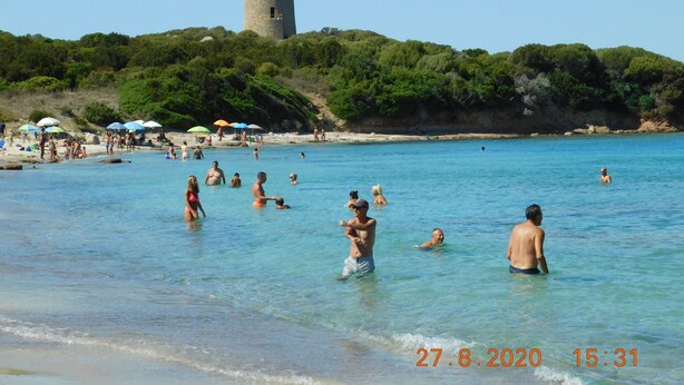 Ein Strand mit badenden und sonnenden Personen, ein Turm im Hintergrund und Sonnenschirme im Sand.